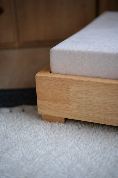 Close-up of a light natural wood bed frame with a white mattress corner resting on a white shag rug.