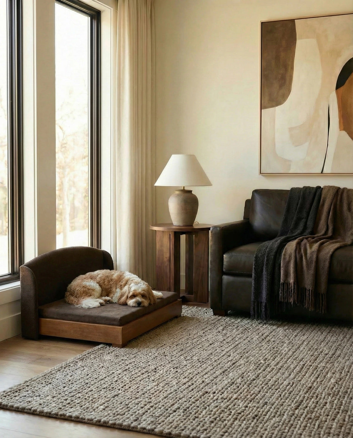 A cozy living room scene with a small brown pet chair and bed on which a fluffy light brown dog is sleeping. A dark leather sofa with a draped throw blanket sits beside a side table holding a table lamp, all on a large woven rug in front of a window with beige curtains.